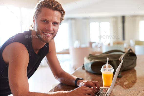 Millennial white man checking fitness app on laptop at home after a workout, smiling to camera