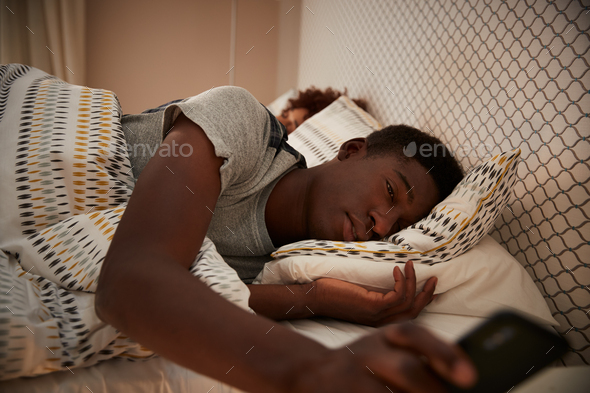 Millennial African American man half asleep in bed holding smartphone
