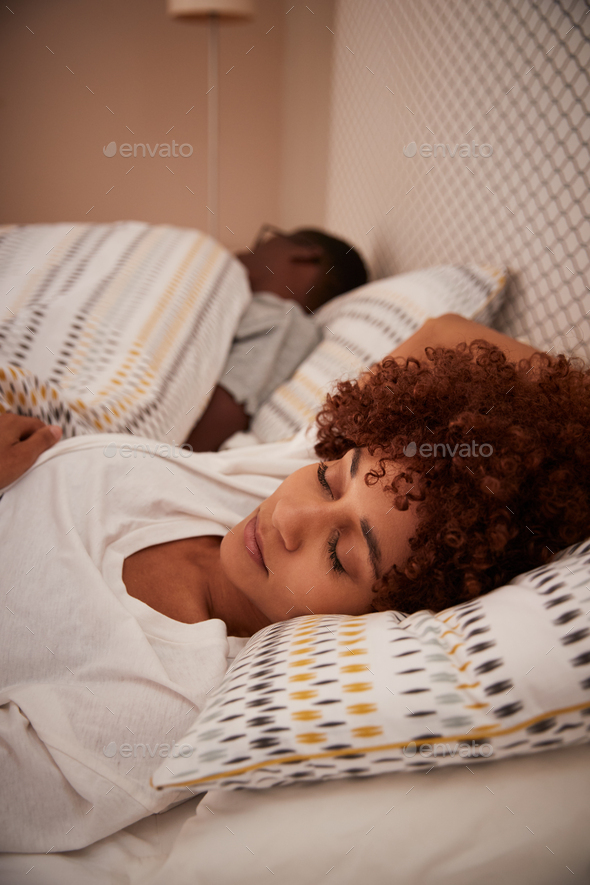 Millennial African American woman lying asleep in bed, her partner in the background, vertical
