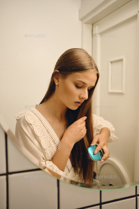 Young romantic girl in white dress dreamily brushing her hair in front of the mirror