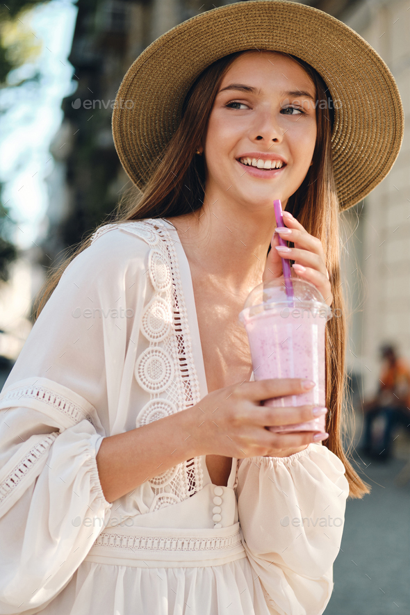 Young attractive smiling girl in white dress and hat holding smoothie joyfully on cozy city street
