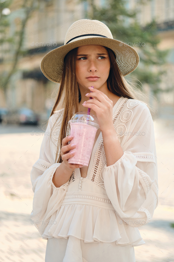 Attractive upset girl in dress and hat holding smoothie sadly looking aside standing on city street