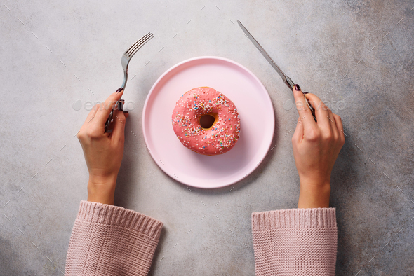 Female hands holding fork and knife cutting donut on pink plate over stone background. Top view - Stock Photo - Images