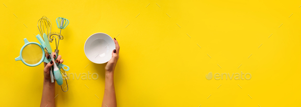 Woman hand holding kitchen utensils on yellow background. Baking tools - bowl, brush, whisk, spatula - Stock Photo - Images