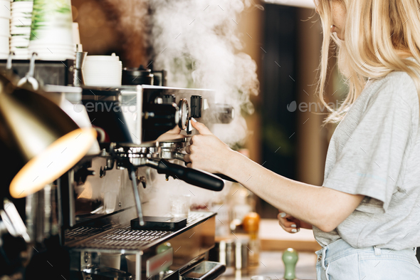 A young pretty thin blonde,dressed in casual outfit,is cooking coffee in a popular coffee shop