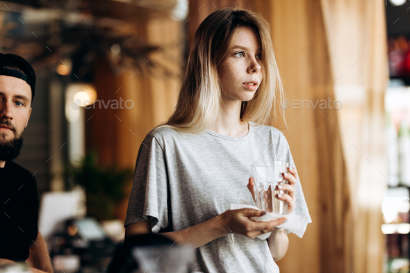 A youthful pretty blonde girl,dressed in casual style, holds a clean glass and looks at the window - Stock Photo - Images
