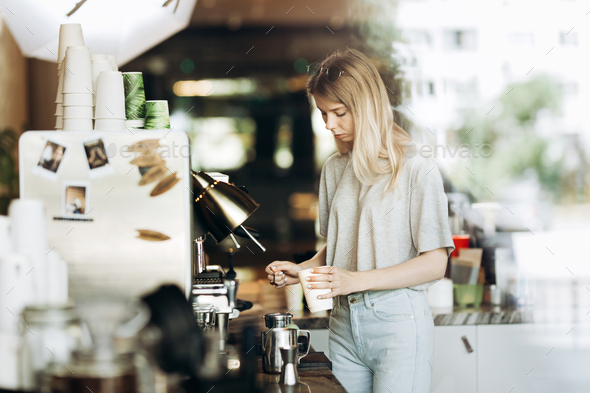 A young pretty thin blonde with long hair,dressed in casual outfit,is cooking coffee in a modern