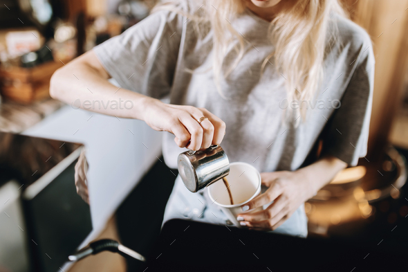 A pretty thin blonde girl with long hair, dressed in casual outfit, pours coffee into a glass in a