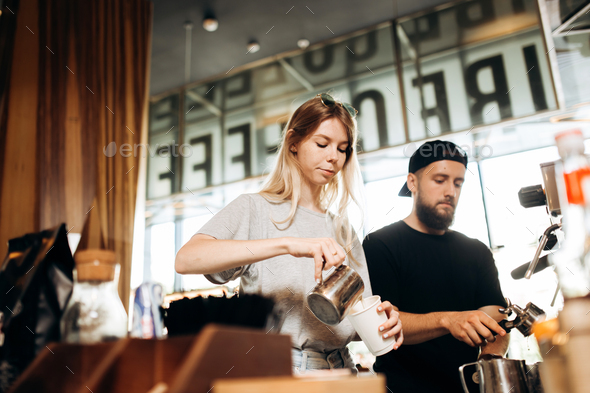 Two young baristas,a blonde girl and stylish man with beard,are shown cooking coffee together in a - Stock Photo - Images