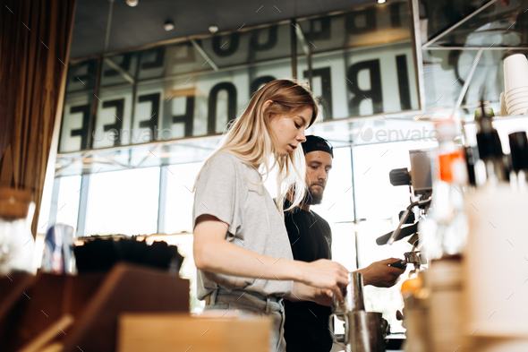 Two young baristas,a blonde girl and stylish man with beard,are shown cooking coffee together in a - Stock Photo - Images
