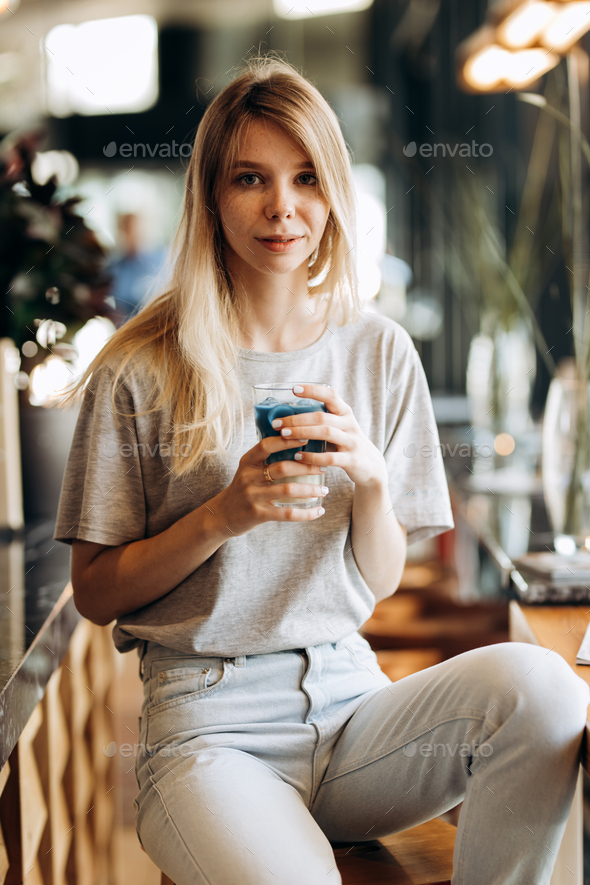 A good looking slim blonde girl,wearing casual style, sits on the chair and holds a cup of coffee in - Stock Photo - Images