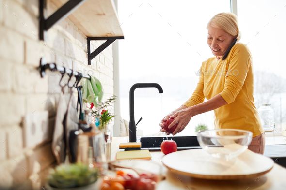 Cheerful woman washing fruit while talking on phone