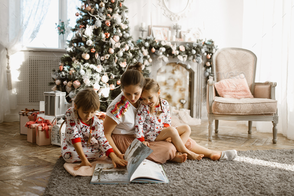 Young beautiful mother with two little daughters sit on the carpet and read book near the New Year - Stock Photo - Images