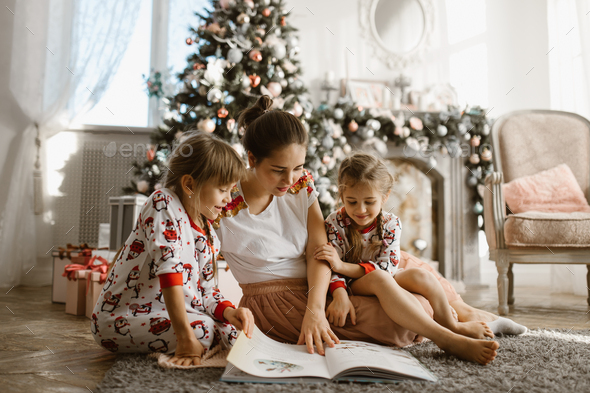 Young beautiful mother with two little daughters sit on the carpet and read book near the New Year - Stock Photo - Images