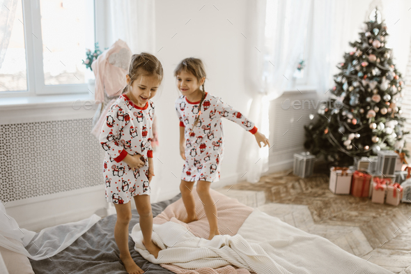 Two charming little girls in their pajamas are having fun jumping on a bed in a sunlit cozy bedroom - Stock Photo - Images