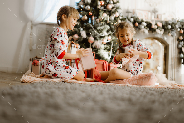 Two happy little sisters in pajamas sit on the carpet and open New Year's gifts in the light - Stock Photo - Images