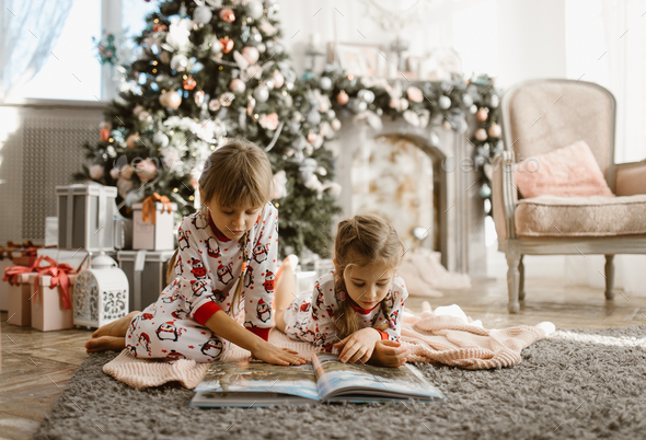 Two little sisters sit on the carpet and read book near the New Year's tree with gifts in the - Stock Photo - Images