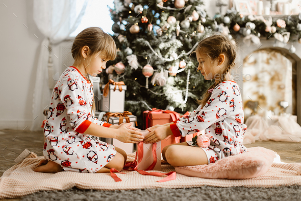 Two little sisters in pajamas sit on the carpet and open New Year's gifts in the light cozy room - Stock Photo - Images