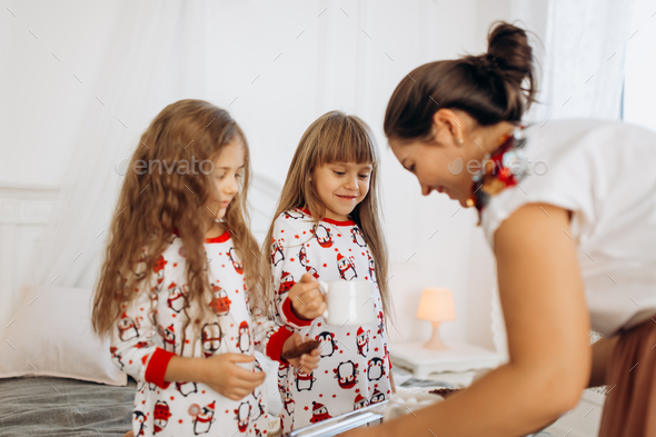 Young mother is bringing cocoa with Marshmallows and cookies to her daughters in pajamas sitting on - Stock Photo - Images