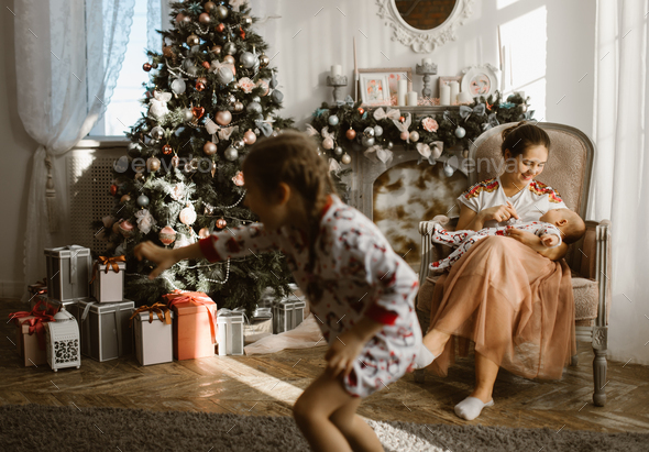 Beautiful mother sits in the armchair with her little baby next to the fireplace and New Year's - Stock Photo - Images