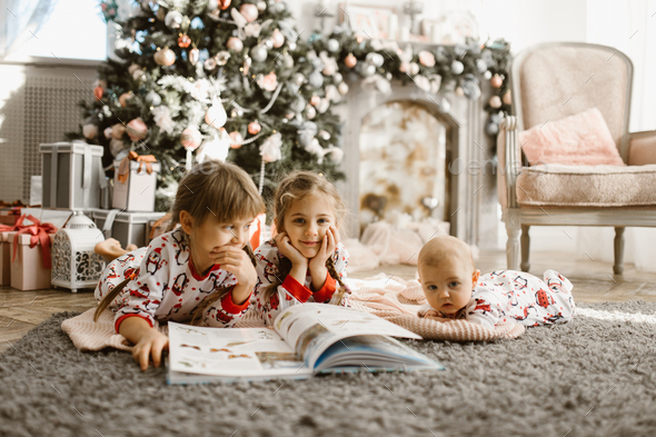 Two little sisters and a tiny brother lie on the carpet and read book near the New Year's tree - Stock Photo - Images