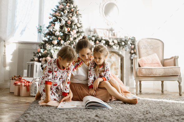 Young beautiful mother with two little daughters sit on the carpet and read book near the New Year - Stock Photo - Images