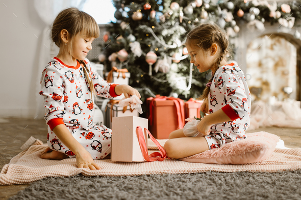Two little sisters in pajamas sit on the carpet and open New Year's gifts in the light cozy room - Stock Photo - Images