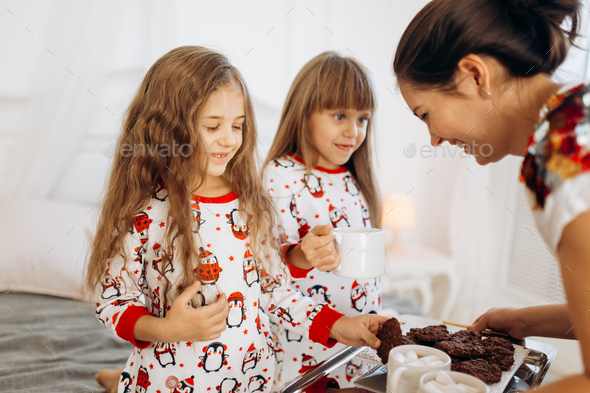 Young mother is bringing cocoa with Marshmallows and cookies to her daughters in pajamas sitting on - Stock Photo - Images
