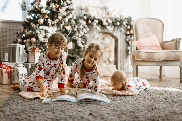 Two little sisters and a tiny brother lie on the carpet and read book near the New Year's tree - Stock Photo - Images