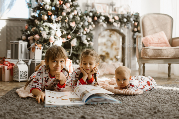 Two little sisters and a tiny brother lie on the carpet and read book near the New Year's tree - Stock Photo - Images