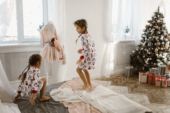 Two charming little girls in their pajamas are having fun jumping on a bed in a sunlit cozy bedroom - Stock Photo - Images