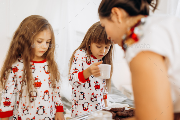 Young mother is bringing cocoa with Marshmallows and cookies to her daughters in pajamas sitting on - Stock Photo - Images