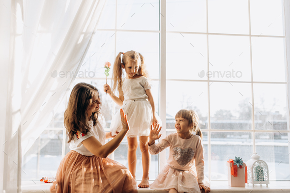 Two little sisters dressed in the beautiful dresses and there young mother sit on the windowsill - Stock Photo - Images