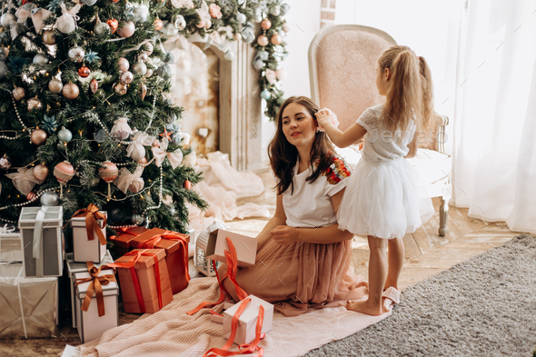 Happy young mother and her little daughter in nice dress sit near the New Year's tree and open - Stock Photo - Images