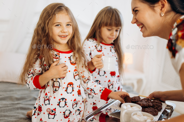Young mother is bringing cocoa with Marshmallows and cookies to her daughters in pajamas sitting on - Stock Photo - Images