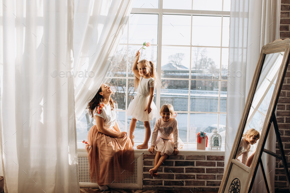 Two little sisters dressed in the beautiful dresses and there young mother sit on the windowsill - Stock Photo - Images