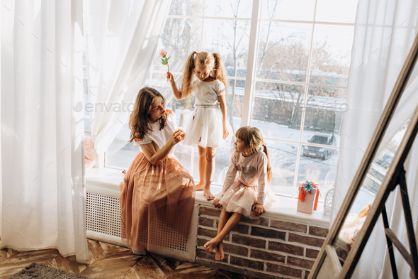 Two little sisters dressed in the beautiful dresses and there young mother sit on the windowsill - Stock Photo - Images