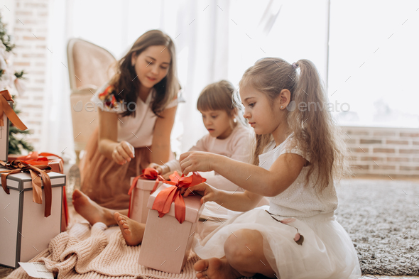 Happy young mother and her two charming daughters in nice dresses sit on the carpet and open New - Stock Photo - Images
