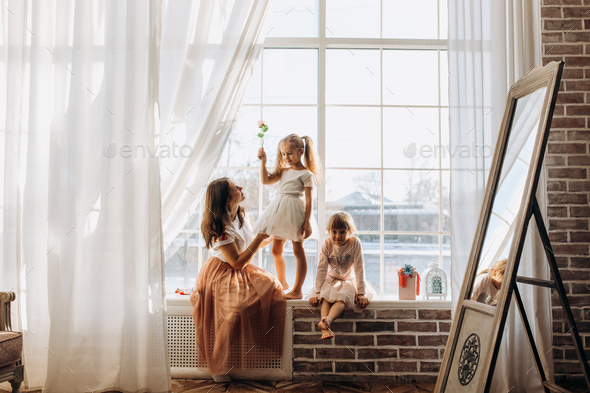 Two little sisters dressed in the beautiful dresses and there young mother sit on the windowsill - Stock Photo - Images