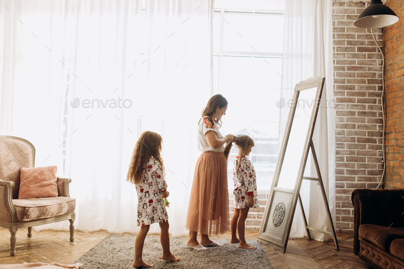 Young mother combing her little daughter's hair standing in front of the mirror and her second - Stock Photo - Images