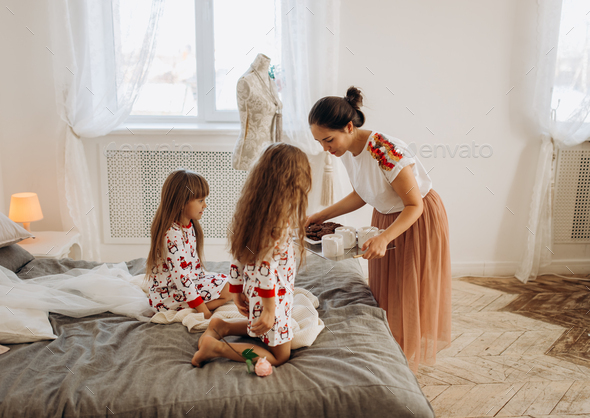Young beautiful mother is bringing cocoa with Marshmallows and cookies to her daughters sitting on - Stock Photo - Images