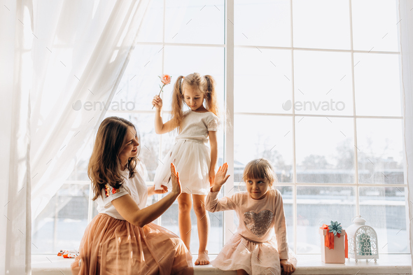 Two little sisters dressed in the beautiful dresses and there young mother sit on the windowsill - Stock Photo - Images