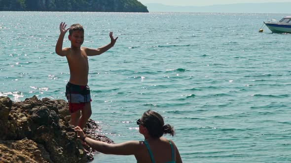 Caucasian kid waves at camera, and jumps into the sea from cape, while his mother watching. Slow mot alt