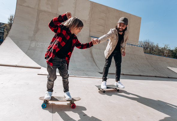 Father and his little son dressed in the casual clothes ride skateboards in a skate park with slides - Stock Photo - Images