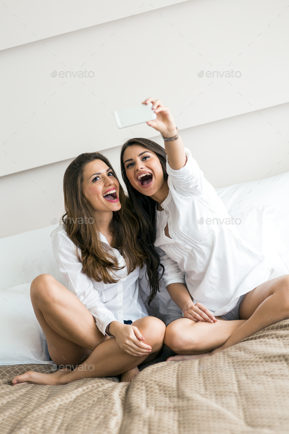 Two hot girls lying on a bed taking a photo of themselves with - Stock Photo - Images