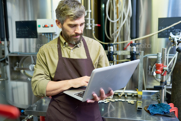 Serious handsome brewer using laptop in factory shop