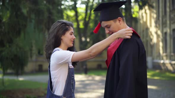 Side View Loving Girlfriend Adjusting Graduation Toga of Boyfriend Gesturing Thumbs Up Smiling As alt