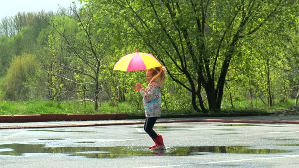Happy funny  child by girl with a multicolored umbrella jumping on puddles in rubber boots
