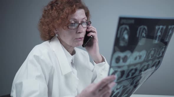 Elderly Caucasian Woman Doctor Analyze Xray Image and Talk with Mobile Phone About Diagnostics in alt