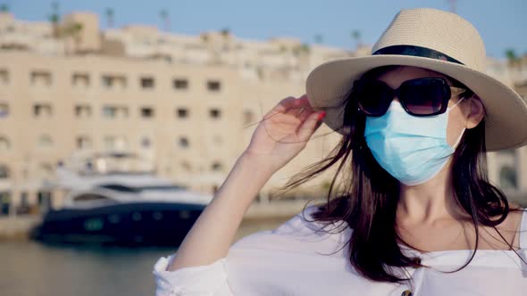 Young Woman in Protective Mask, Summer Clothes, Sunglasses and Sun Hat, on the Background of a Yacht alt